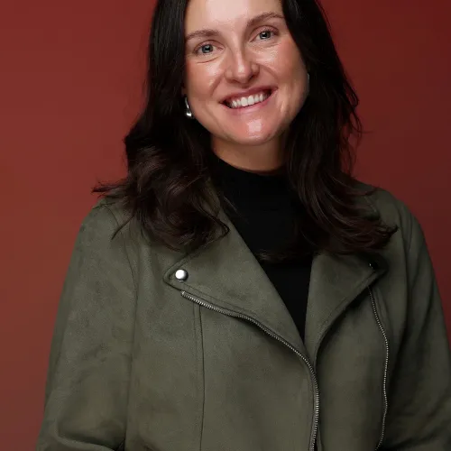 Smiling woman with dark hair wearing a green jacket and black top against a reddish brown background