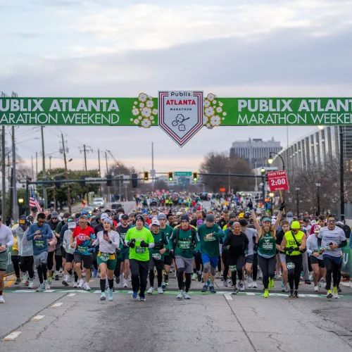 Runners start the Publix Atlanta Marathon Weekend race under a green banner on city street at dawn.