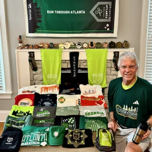 Man sitting beside table with Atlanta marathon shirts, medals, towels, and running gear in a home setting.