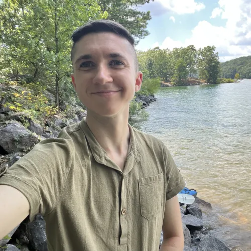 Smiling young man takes selfie by a rocky lakeside with trees and cloudy sky in the background