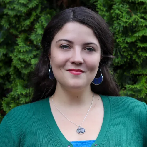 Portrait of a smiling woman wearing green sweater, silver tree pendant necklace, and blue earrings against green foliage.