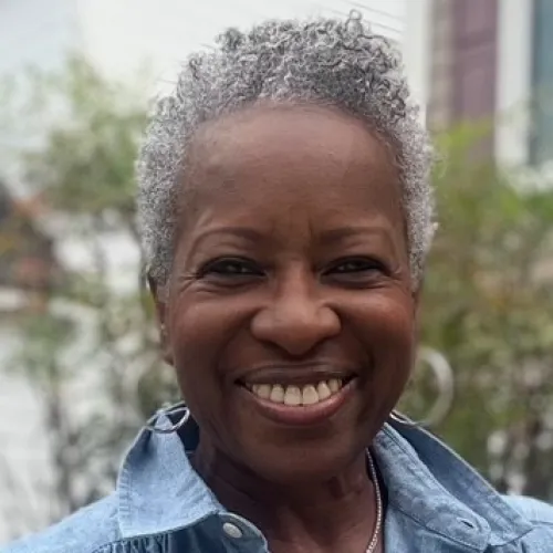 Smiling older woman with short curly gray hair wearing a blue button-up shirt and layered necklaces outdoors.