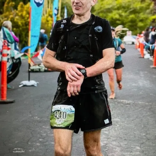 Runner in black sportswear and yellow cap checks watch during an outdoor race with event flags and spectators in background.