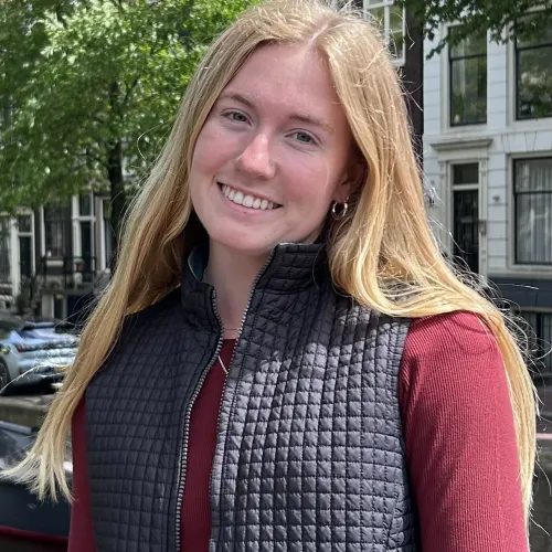Young woman with long blonde hair smiling outdoors in a black quilted vest and maroon shirt near a canal.