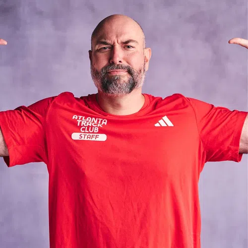 Bearded man in red Atlanta Track Club staff shirt pointing to himself with confident expression.