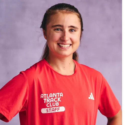 Smiling woman wearing a red Atlanta Track Club Staff shirt with a purple background.