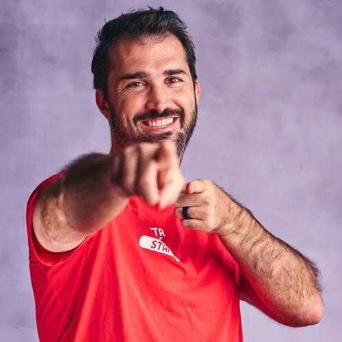 Smiling man with beard wearing red shirt pointing directly at the camera against a blurred purple background.