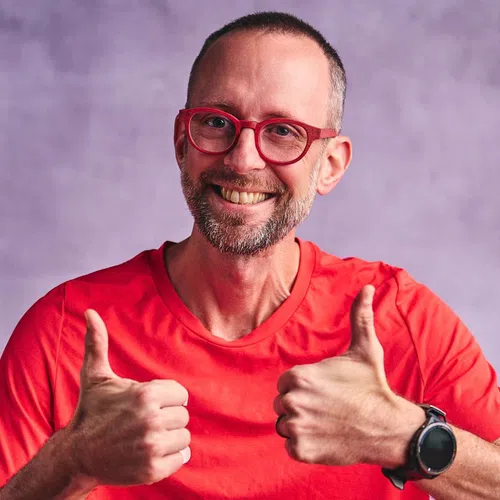 Smiling man with glasses and beard wearing red shirt showing double thumbs up against purple background