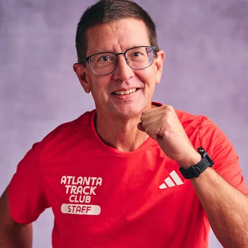 Smiling man in glasses wearing a red Atlanta Track Club staff shirt with a fist raised in front of a purple background
