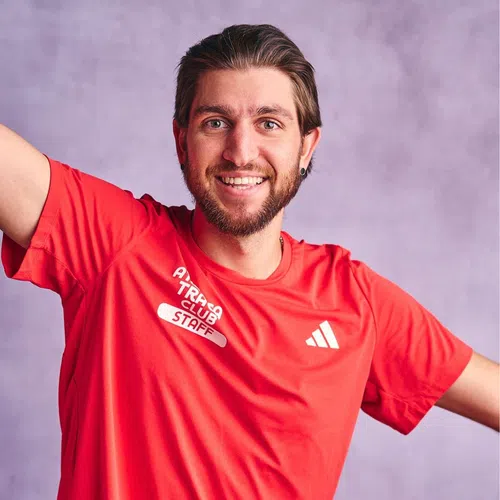 Smiling man in red Adidas staff t-shirt posing with arms raised against a soft purple background