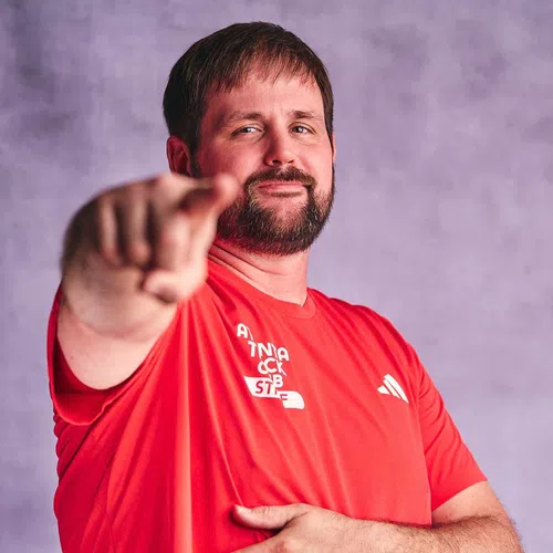 Man with beard pointing forward wearing a red Adidas shirt against a plain purple background
