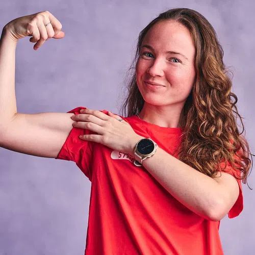 Smiling woman in a red shirt flexes her arm muscle, showing strength and confidence against a purple background.