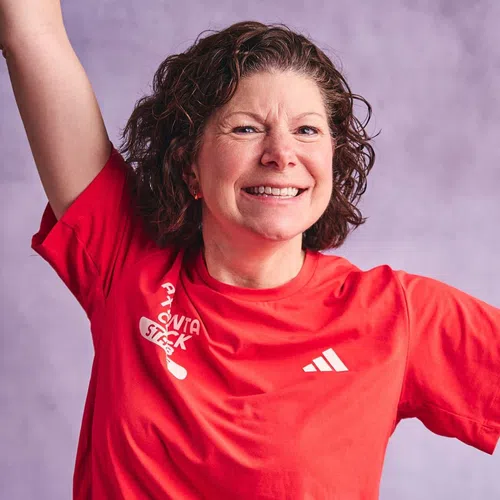 Smiling woman in red Adidas shirt raises arm against purple background, showing enthusiasm and energy.
