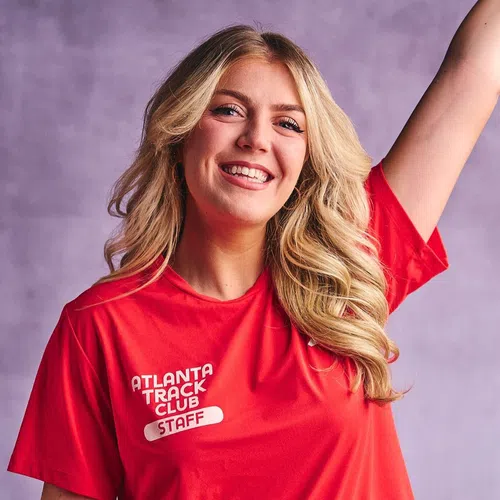 Smiling young woman in red Atlanta Track Club Staff t-shirt raising her arm against a purple background