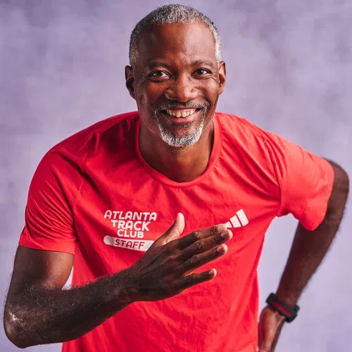 Smiling middle-aged man in red Atlanta Track Club staff shirt posing in a running stance against a purple background