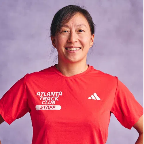 Smiling woman wearing a red Atlanta Track Club staff shirt against a light purple background