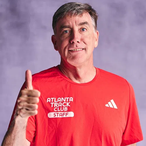 Middle-aged man in red Atlanta Track Club staff shirt smiling and giving thumbs up against purple background