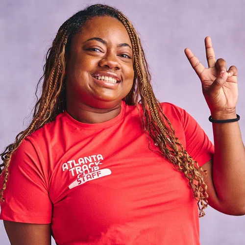 Smiling woman wearing red Atlanta Track Club staff shirt showing peace sign on purple background