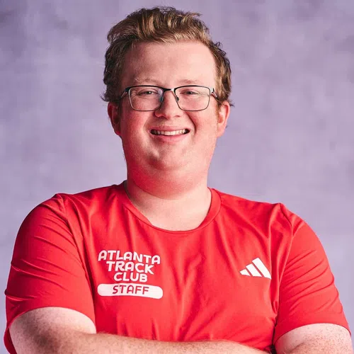Smiling man in glasses wearing a red Atlanta Track Club staff shirt with folded arms against a purple background