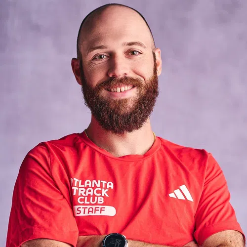 Smiling bald man with a beard wearing a red Atlanta Track Club staff shirt against a purple background