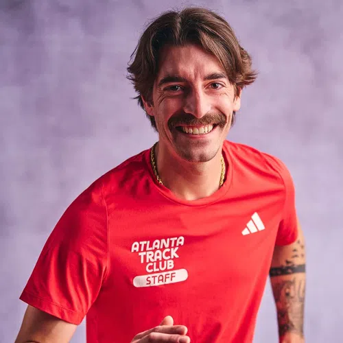 Smiling man with mustache in red Atlanta Track Club staff shirt against purple background