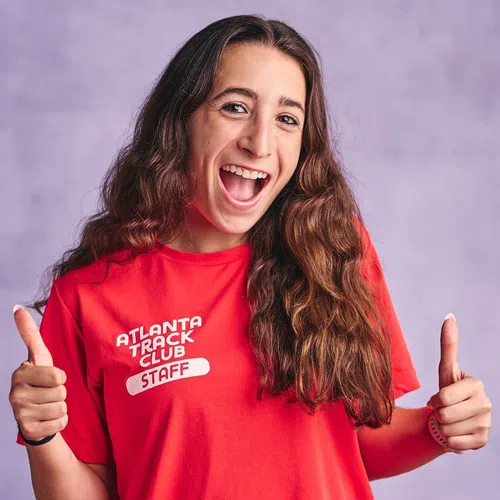 Smiling young woman in red Atlanta Track Club Staff t-shirt giving two thumbs up against a purple background