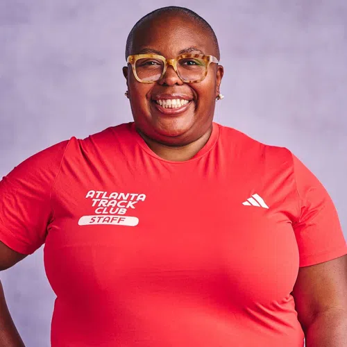 Smiling person wearing large tortoiseshell glasses and red Atlanta Track Club staff shirt on purple background