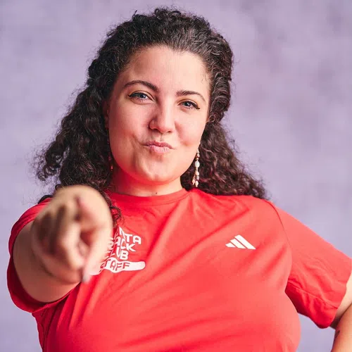 Woman with curly hair in red shirt pointing forward with confident expression against soft purple background