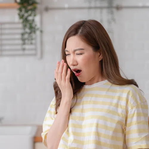 Young woman in striped shirt covering her mouth while yawning in a bright kitchen setting