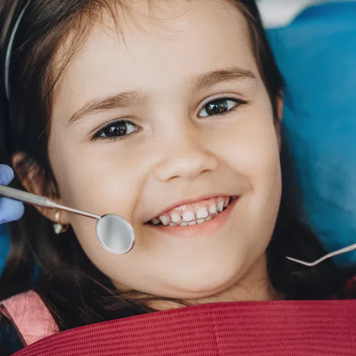 Smiling young girl sitting in dental chair with dentist's hands holding dental tools near her mouth