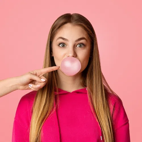 Young woman blowing a pink bubblegum bubble with a finger about to pop it against a pink background.