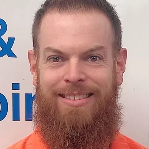 Smiling man with a red beard and styled hair wearing an orange shirt against a white background with blue text.