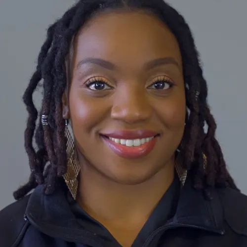 Smiling woman with braided hair wearing black jacket with a blue and white circular logo on gray background.