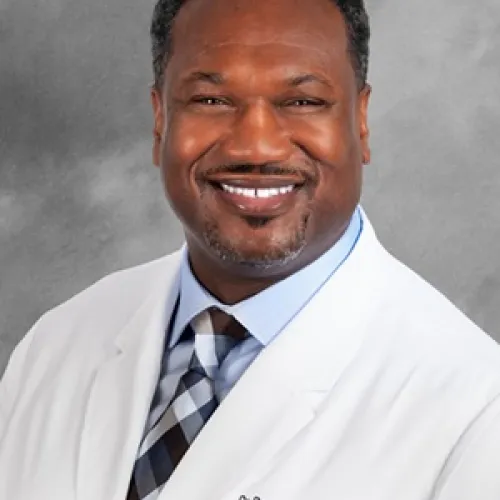 Smiling doctor wearing a white coat and tie, against a gray background.
