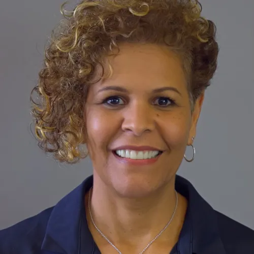 Smiling woman with curly hair wearing a navy shirt and silver cross necklace against a plain gray background