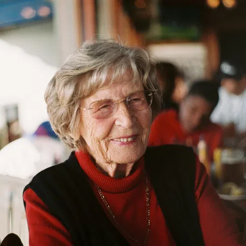 Smiling elderly woman wearing glasses and a red turtleneck sweater sitting in a busy indoor setting.