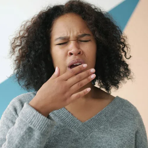 Woman with curly hair yawning with hand covering mouth against colorful geometric wall background