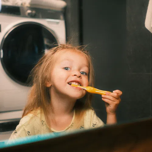 Young child brushing teeth in bathroom with washing machine in the background looking at mirror.