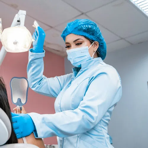 Female dentist in protective gear adjusting dental light while patient holds mirror at dental clinic