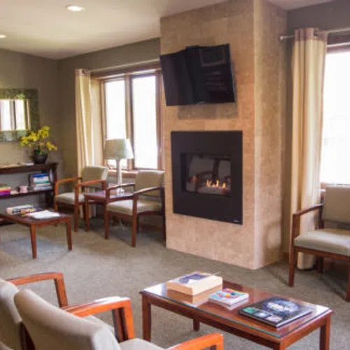 Cozy waiting room with cushioned chairs, a fireplace, flat-screen TV, and natural light from large windows.