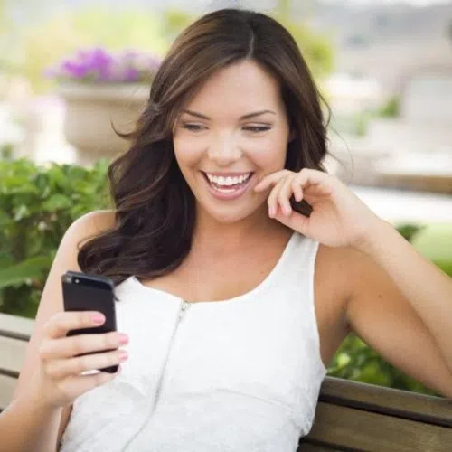 Happy young woman smiling while looking at her smartphone outdoors on a park bench with greenery background