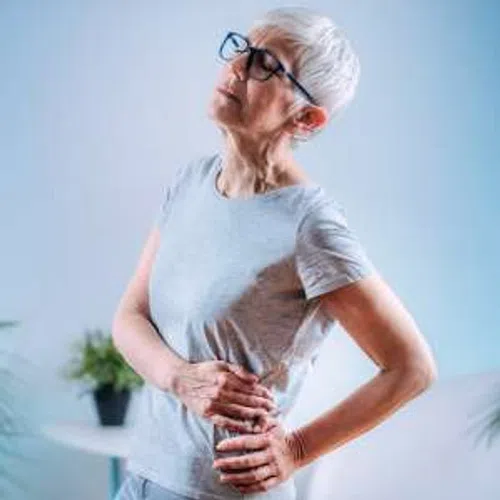 Senior woman with glasses holding her side, expressing pain or discomfort indoors with a blurred plant background