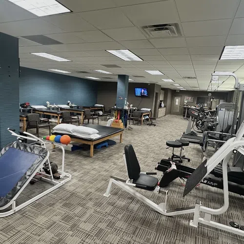 Spacious physical therapy clinic with exercise machines, therapy tables, and rehab equipment in neutral tones.