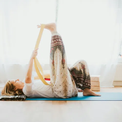 Woman practicing yoga indoors on a mat using a strap to stretch her leg in a bright, peaceful room.