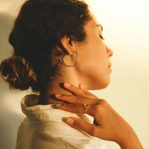 Side profile of a woman with hair in a bun wearing hoop earrings and touching her neck with a ring on her finger.