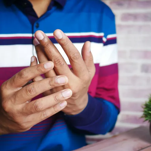 Man in a striped blue shirt rubbing fingers, possibly experiencing joint pain, sitting at a wooden table with a small plant.
