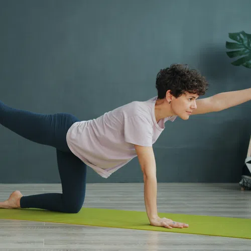 Woman practicing yoga balancing on one hand and leg on green mat indoors with plants in the background