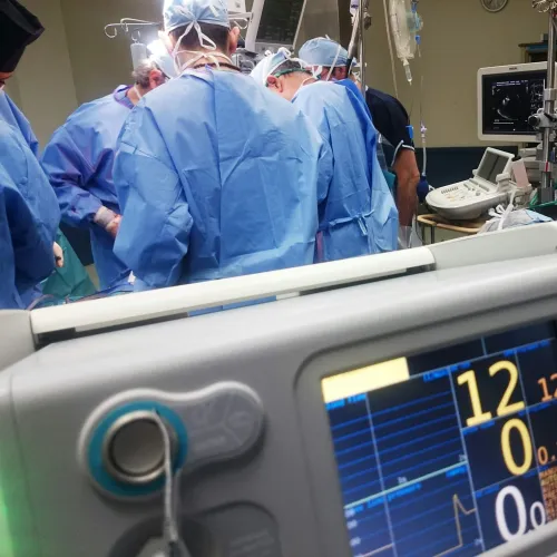 Vital signs monitor in foreground during surgery with medical team in blue scrubs and masks in operating room.