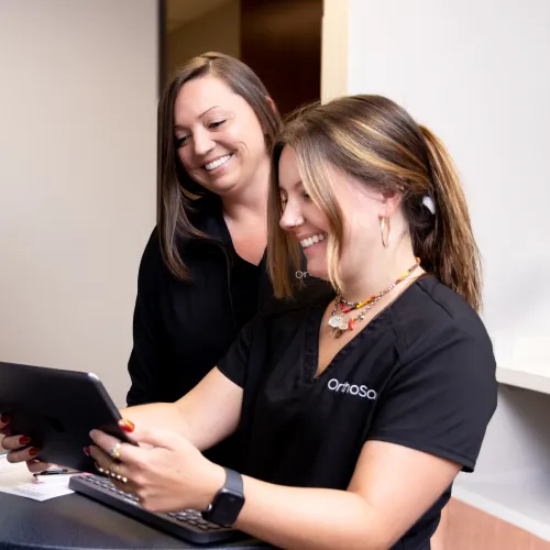 Two female healthcare professionals smiling and reviewing information on a tablet in an office setting.