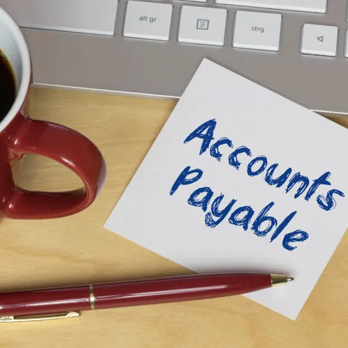 A cup of coffee next to a notepad with 'Accounts Payable' written on it beside a keyboard.